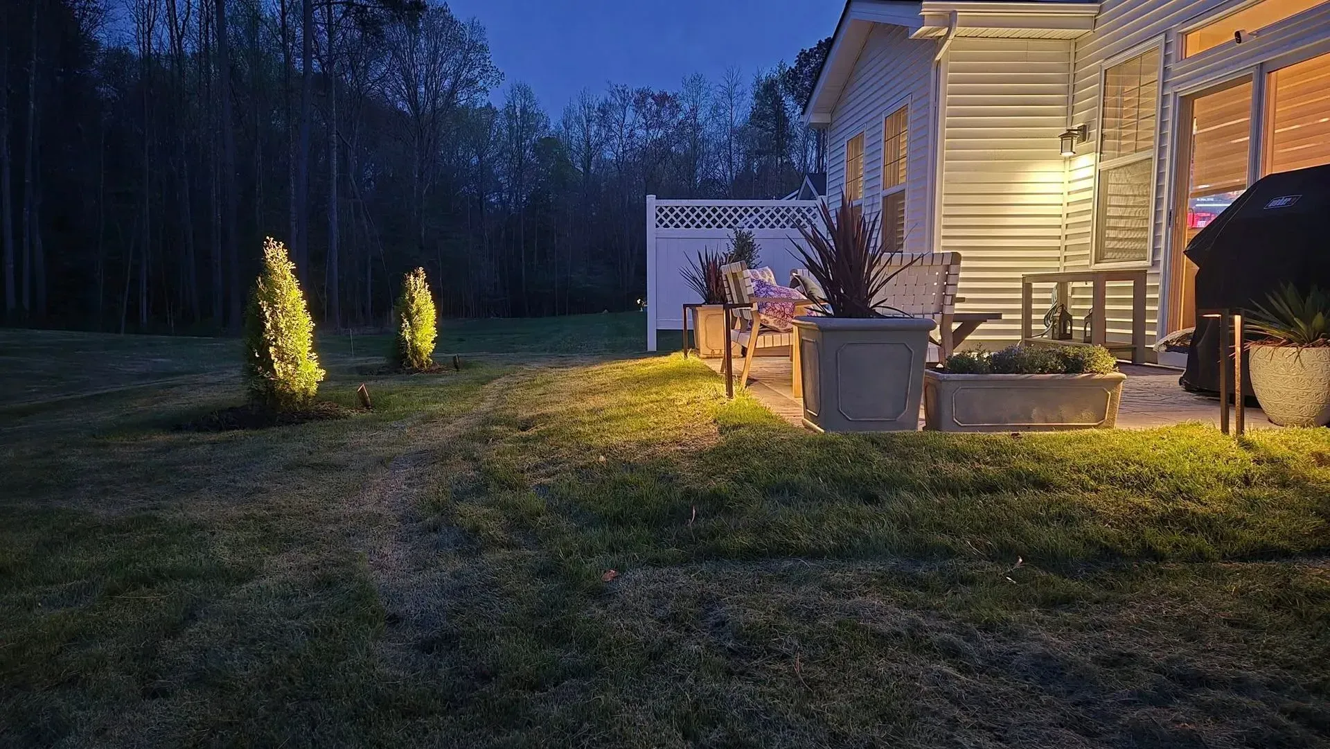 Backyard at dusk with lit yellow shrubs, a patio chair, and a house porch glowing under warm lights