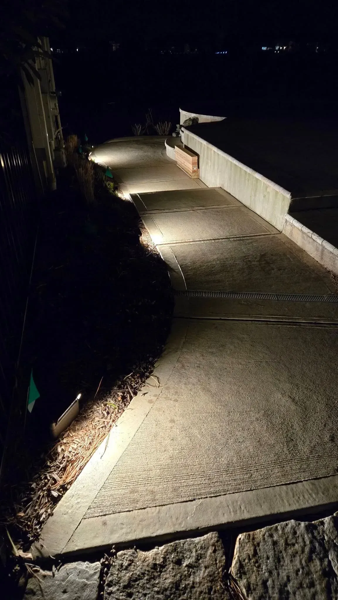 Lit nighttime walkway beside dark water, bordered by a low wall and a flag in the distance.