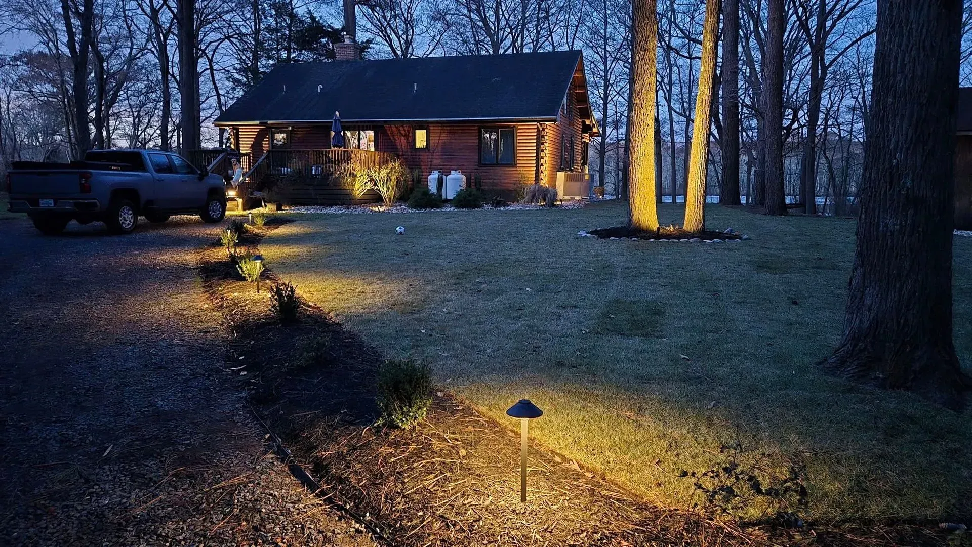 Lit cabin in a wooded yard at dusk, with warm lights along a path and a parked vehicle nearby