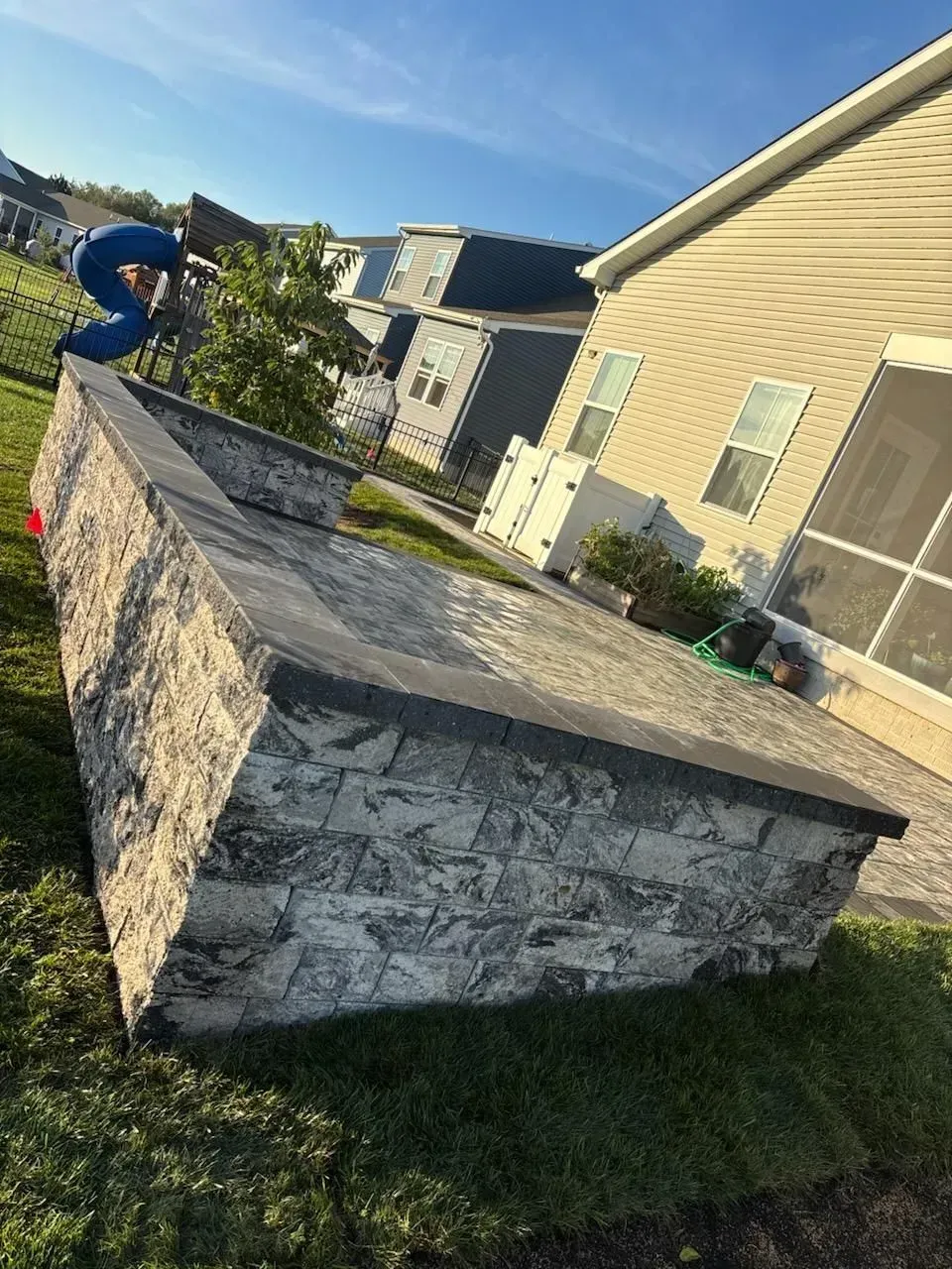 Person crouching on a stone wall in a sunny suburban yard with houses nearby