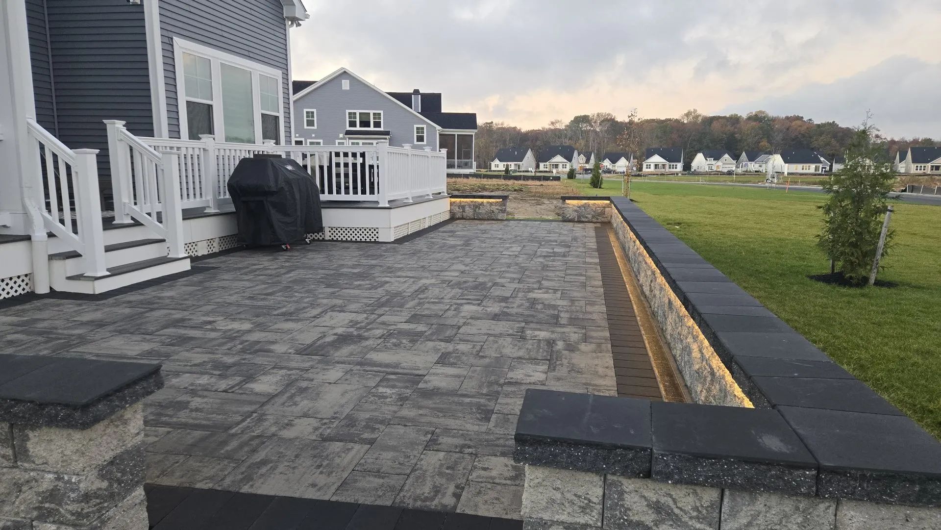 Stone patio beside a house, with white deck railings and a grassy yard at dusk.