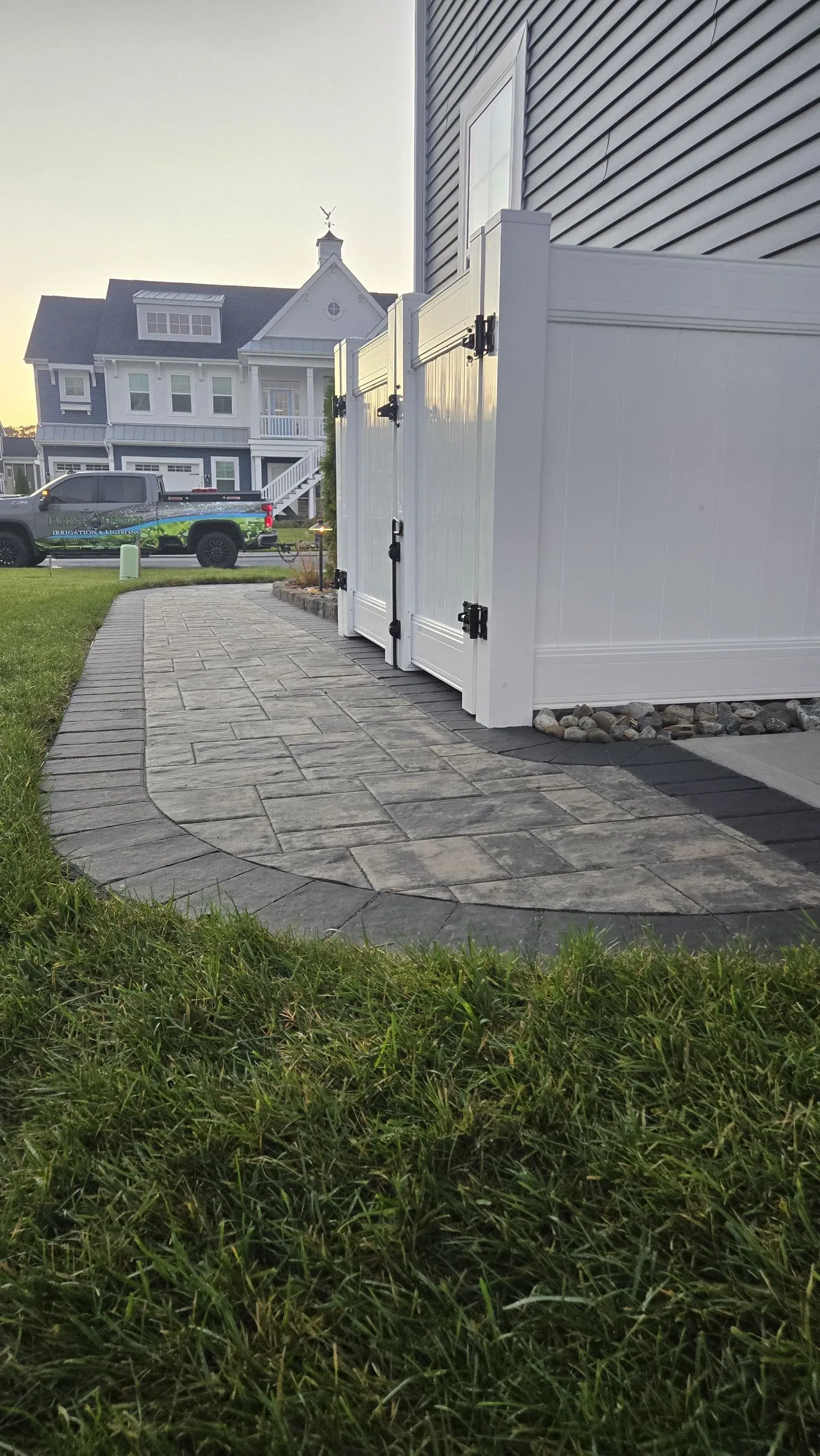 Curved stone walkway beside a white house with patio doors, leading past a grassy yard to neighboring homes.