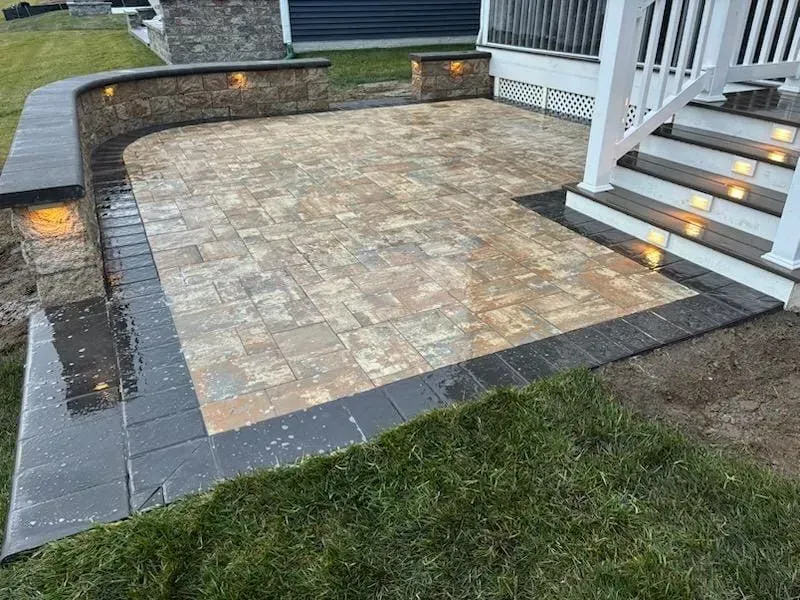 Stone patio with dark pavers, built-in steps, and soft landscape lighting beside a deck.