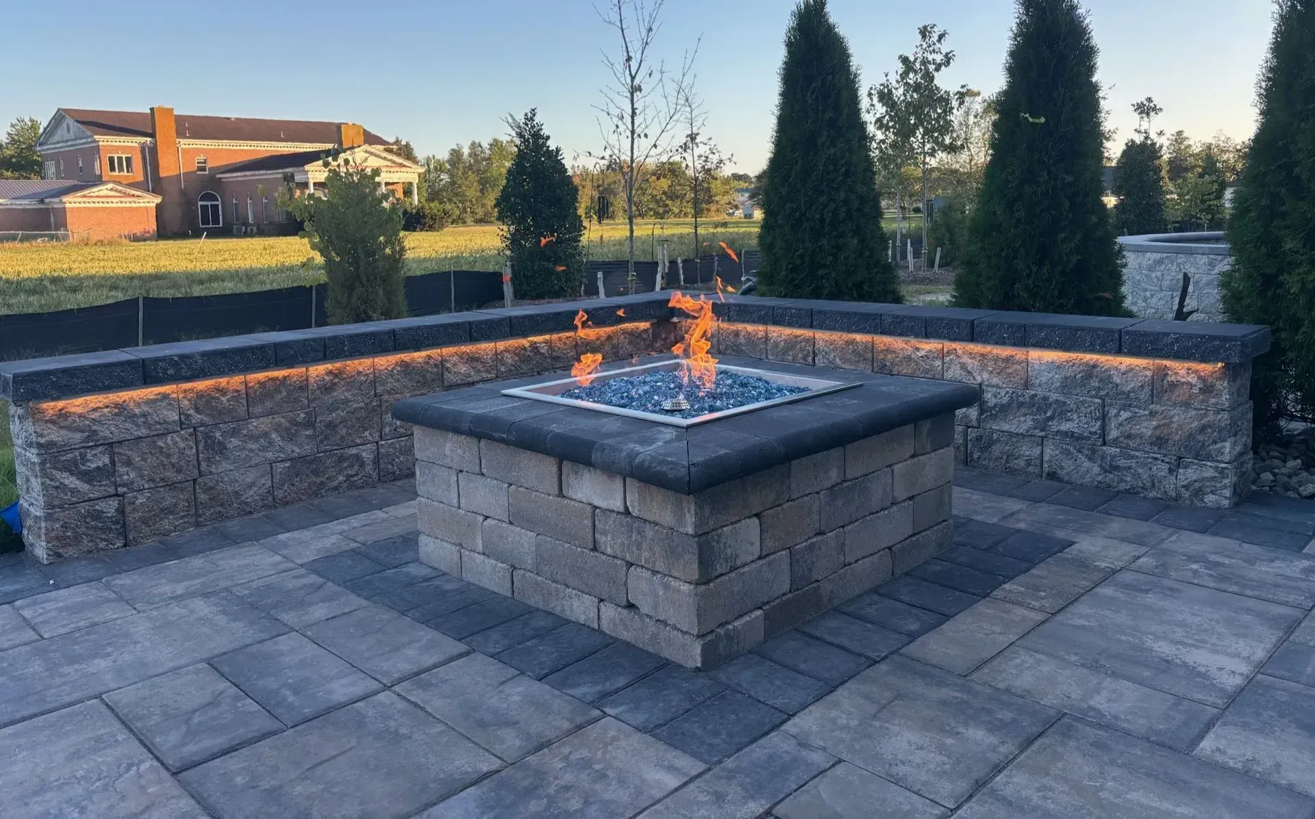 Stone fire pit with flames in a paved backyard patio at sunset, bordered by low stone walls and trees