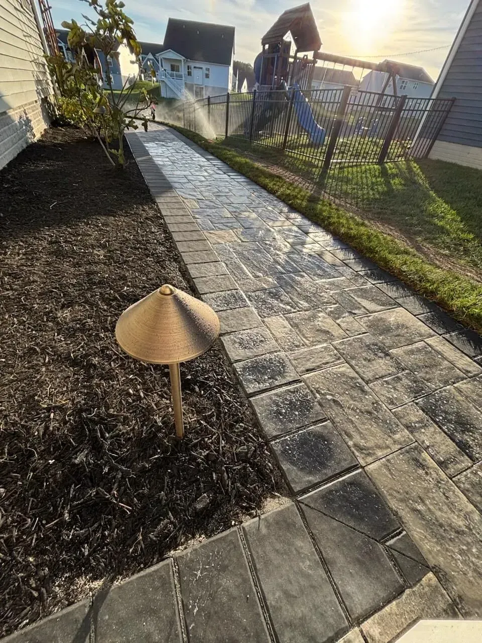 Stone garden path beside mulch beds and a lattice fence, with houses and sunset light in the background