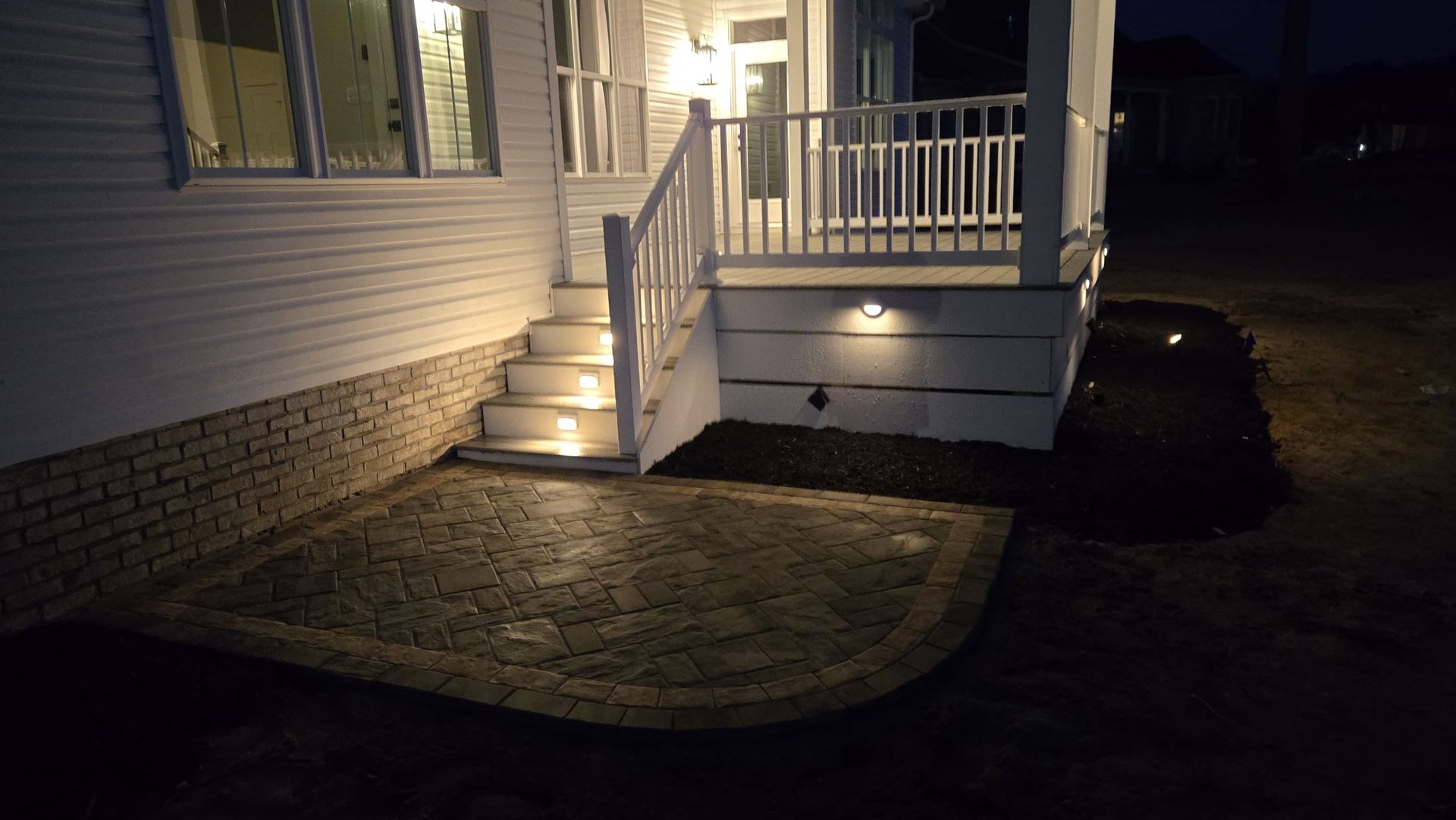 Lit house porch with white railings and steps, beside a curved brick patio at night
