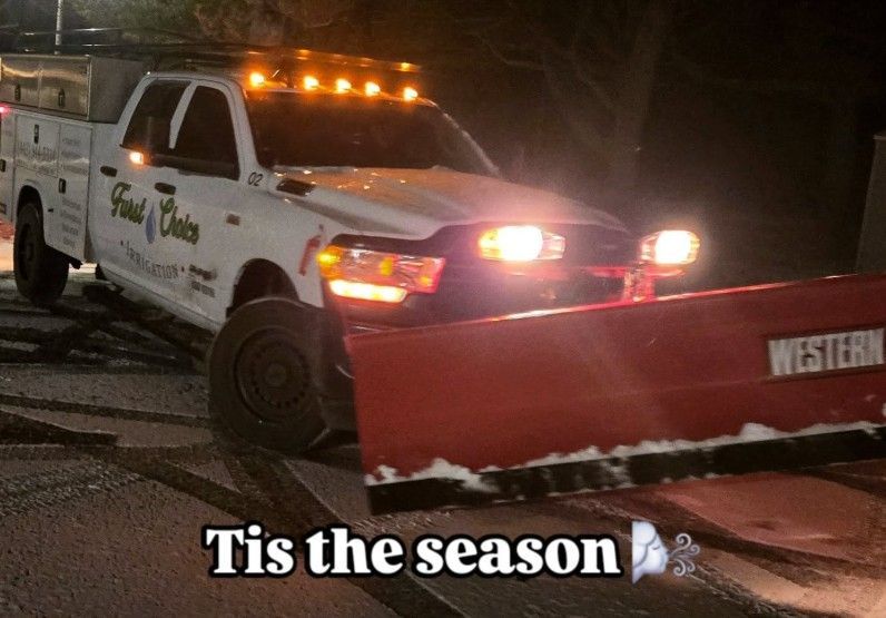 Tow truck with flashing lights pushing a red snowplow at night, captioned “Tis the season”