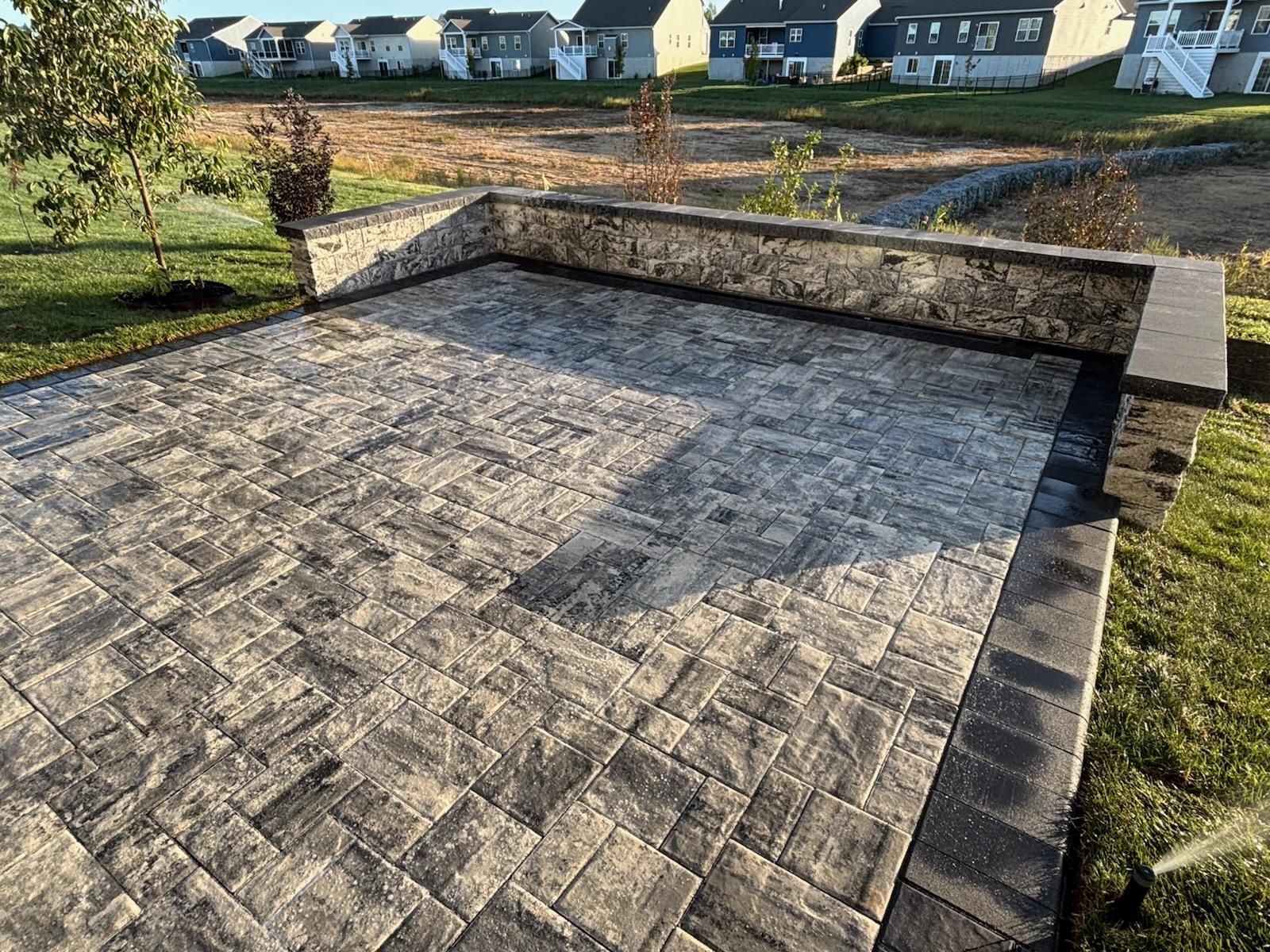 Raised stone patio with a low retaining wall in a grassy yard, suburban houses in the background