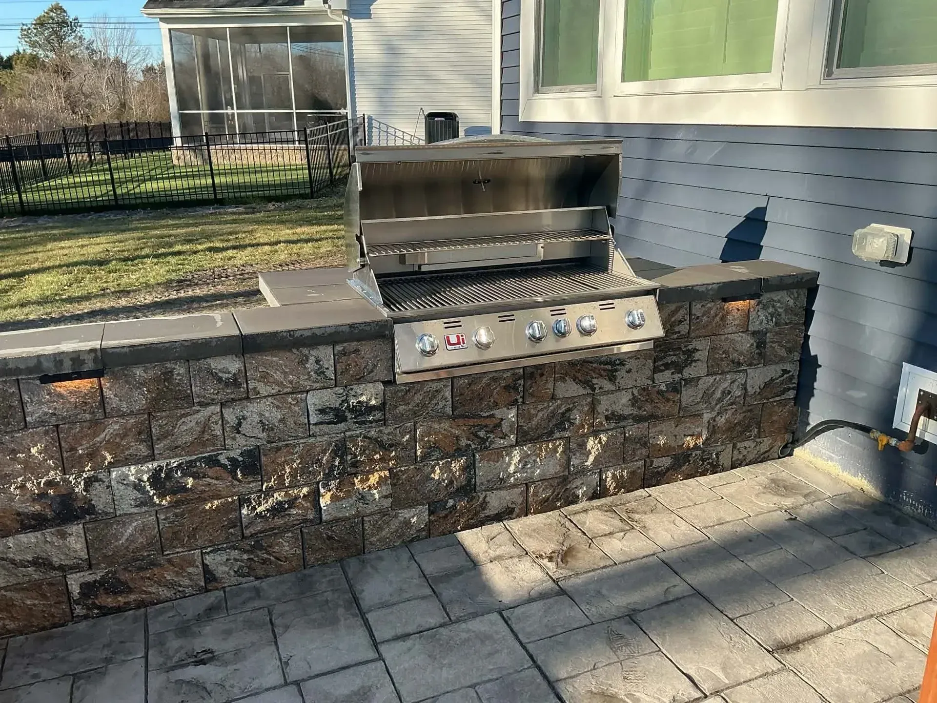 Stone outdoor grill built into a low wall beside a house patio with gray pavers.