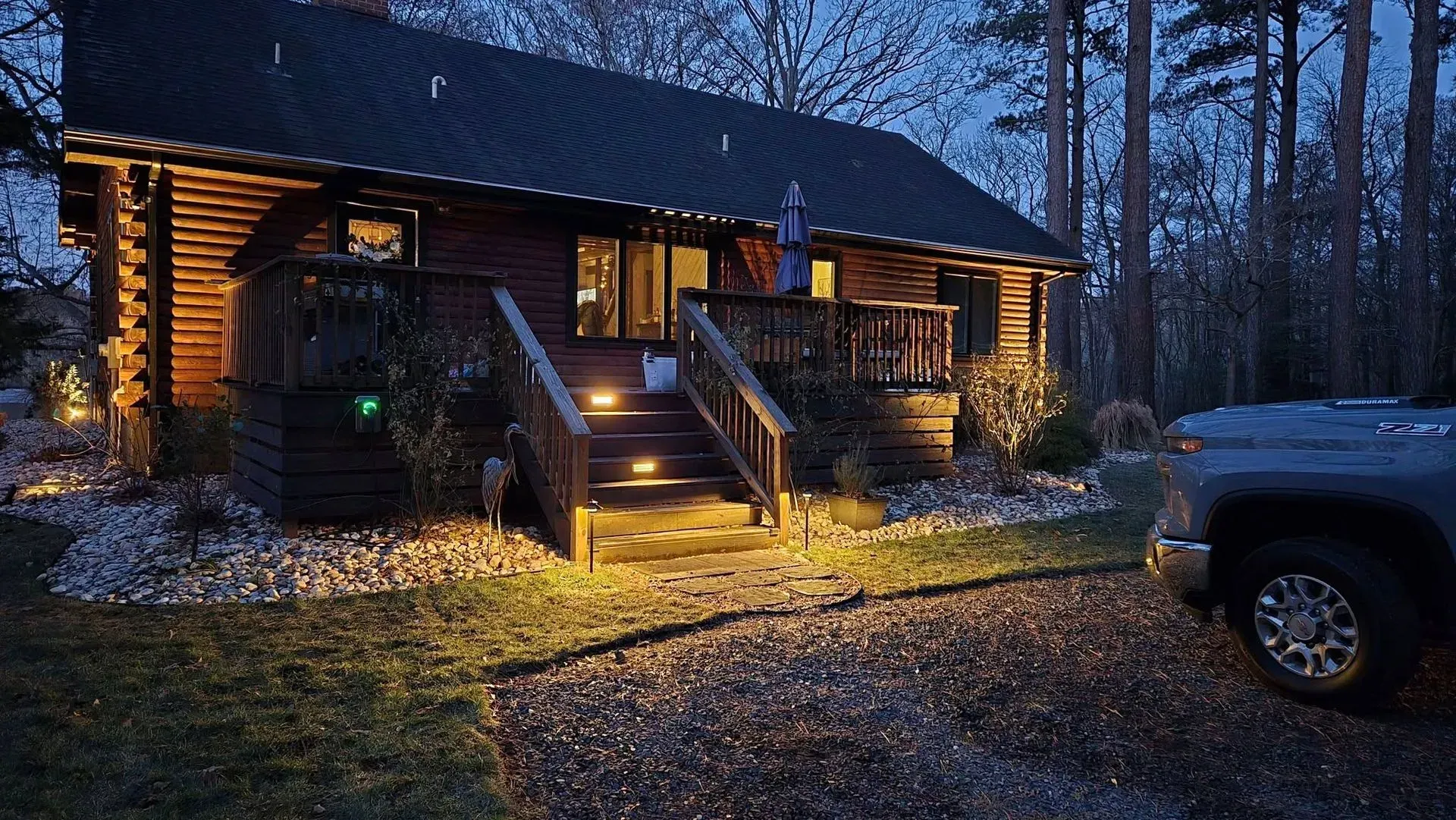 Cozy log cabin at dusk with lit porch steps and a parked truck in a wooded yard
