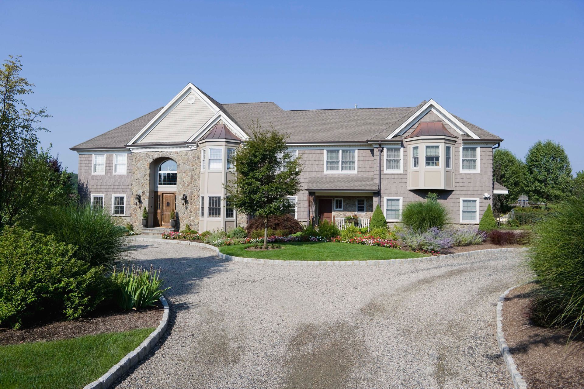 Large beige suburban house with a gravel driveway, landscaped garden, and trees in front.