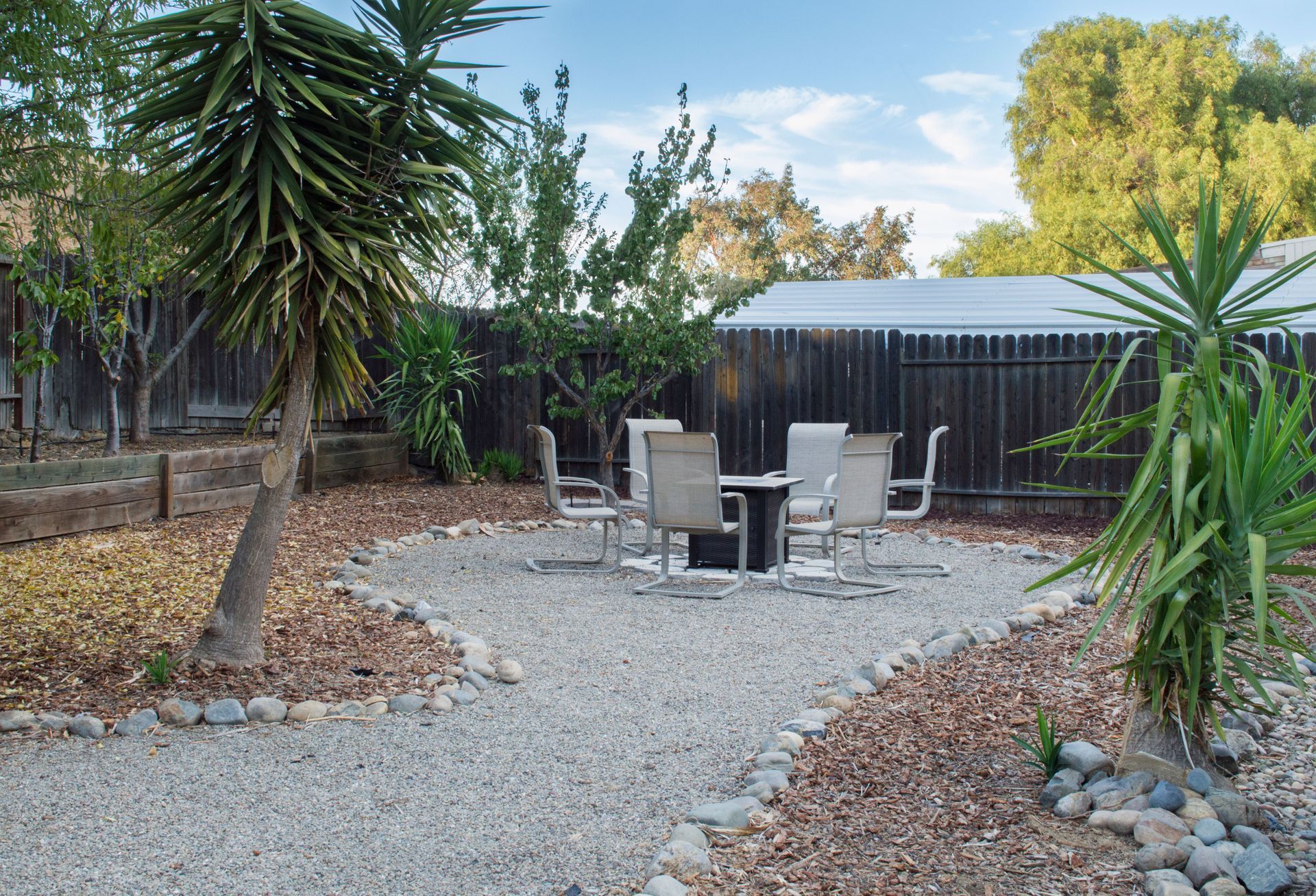 Backyard patio with gravel path, outdoor chairs, and tropical plants near a dark fence
