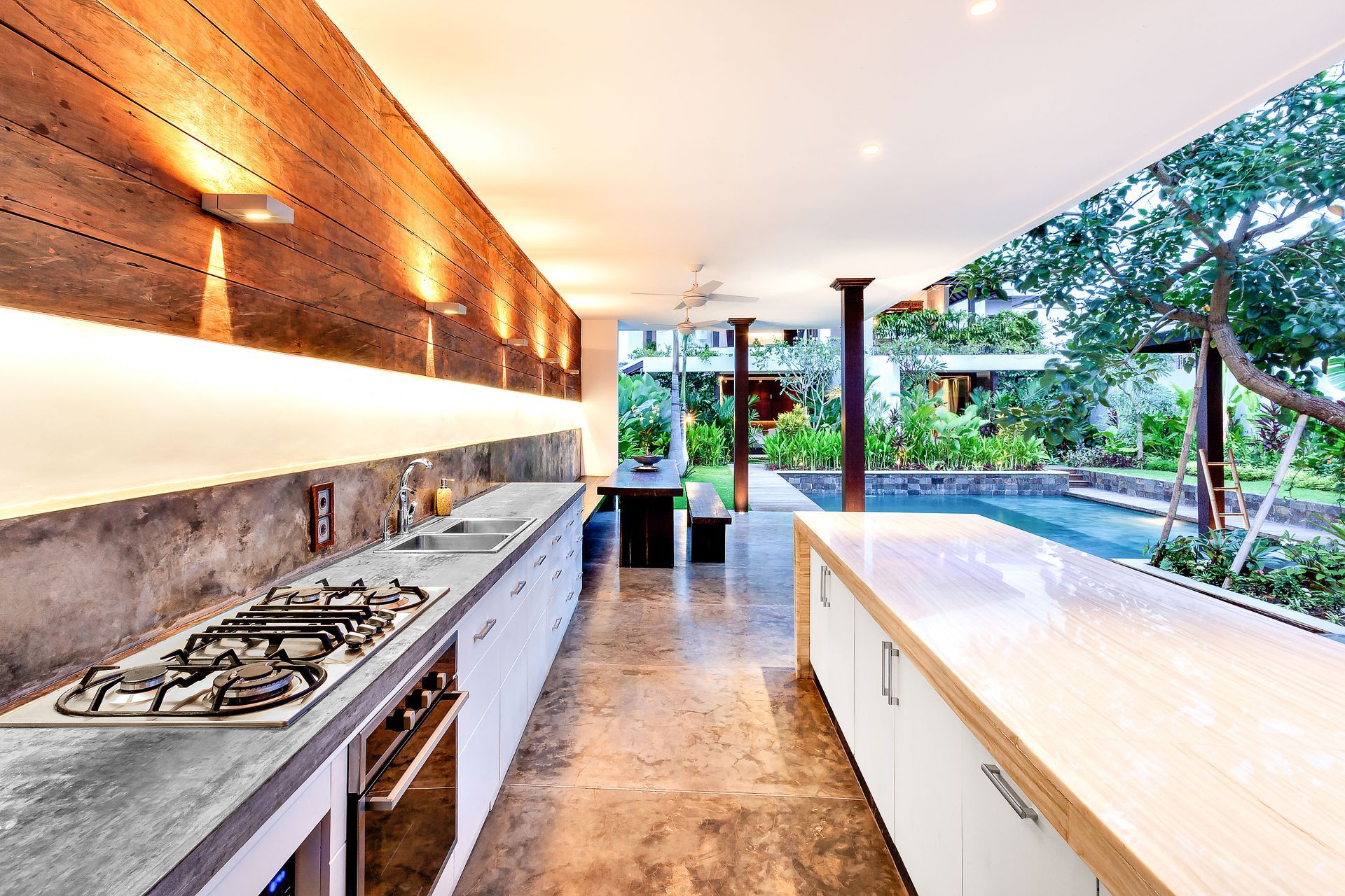 Modern outdoor kitchen with stainless counters, wood accents, and a view of a pool and garden.
