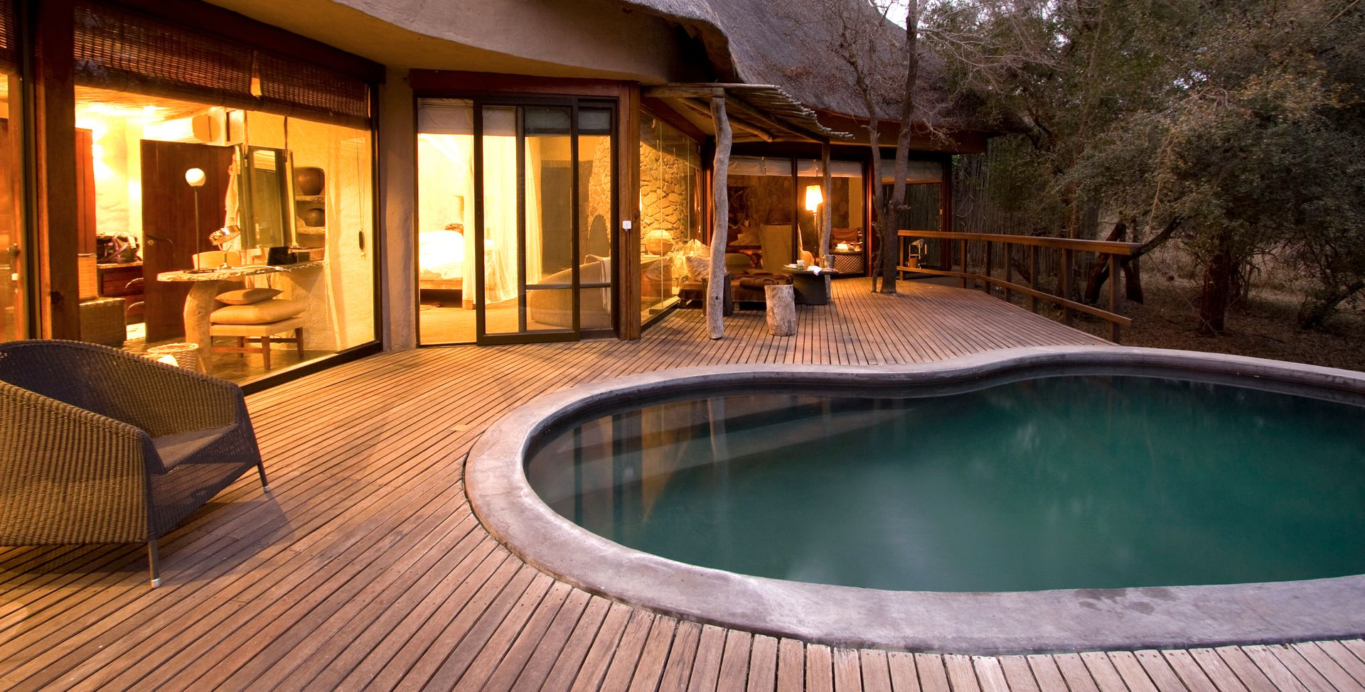 Wooden deck with a curved outdoor pool beside a warmly lit modern house at dusk