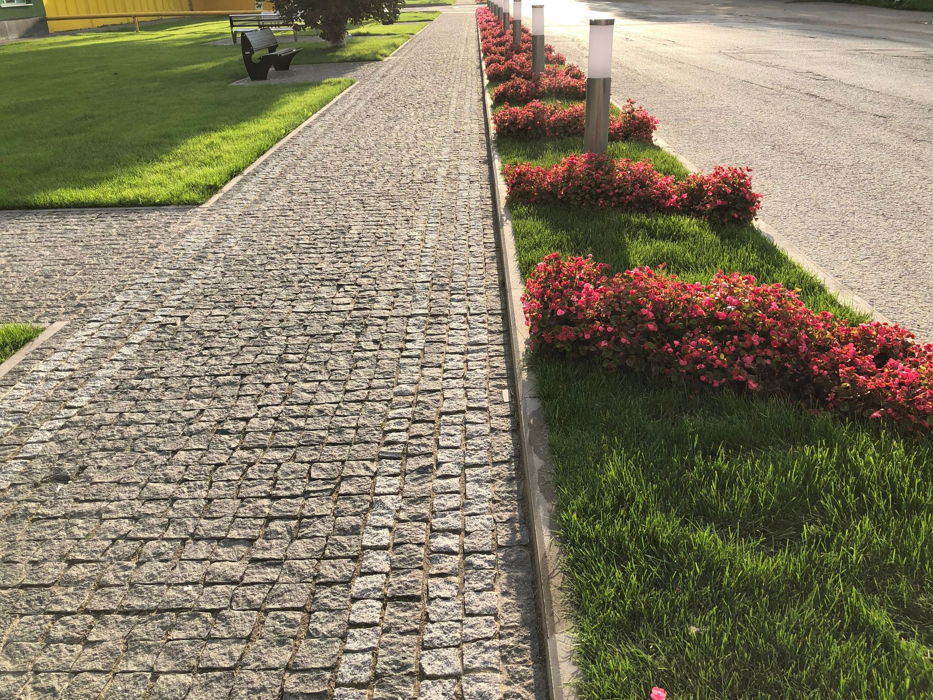 Cobblestone walkway beside a lawn and red flowers along a curb near a road