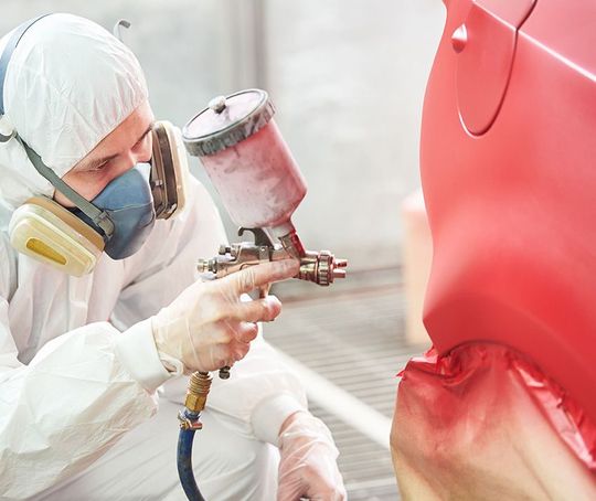 A man in a protective suit is spray painting a red car.