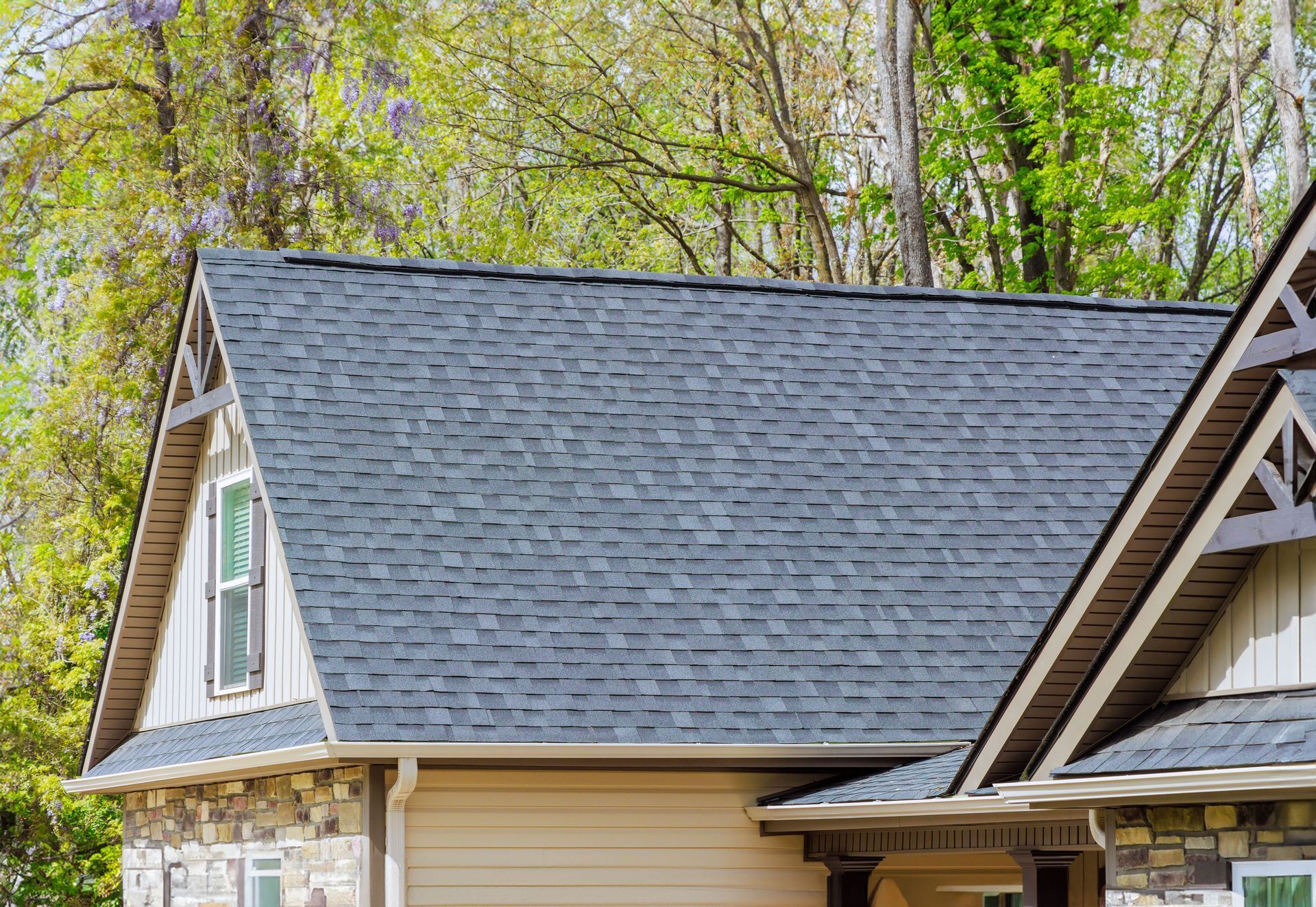 Gray asphalt shingle roof on a house, with a window and a wooded background.