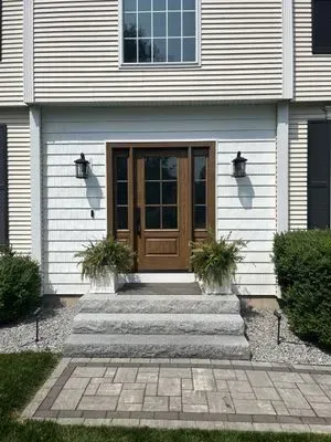 Exterior of a house with a brown door, sidelights, and gray stone steps. Two potted plants flank the door.