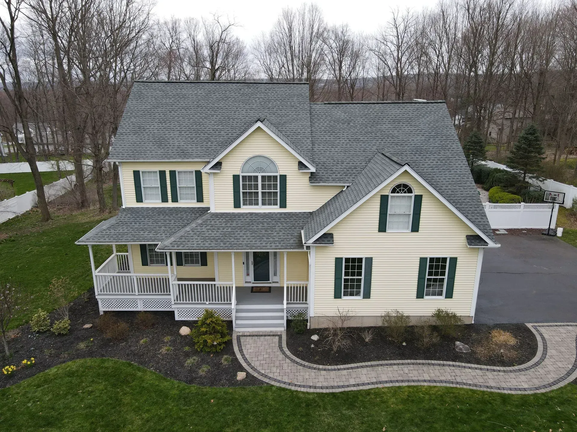 A two-story, yellow house with green shutters, a covered porch, and a curved stone walkway in a yard with bare trees.