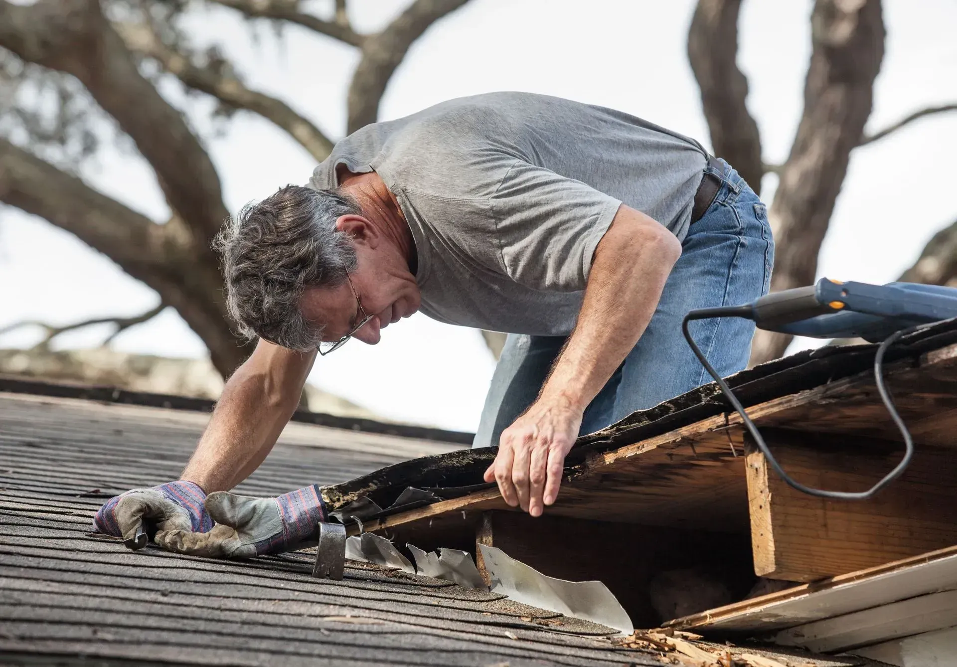 A worker wearing gloves kneels on a roof, repairing a section of exposed wooden deck near the eaves.