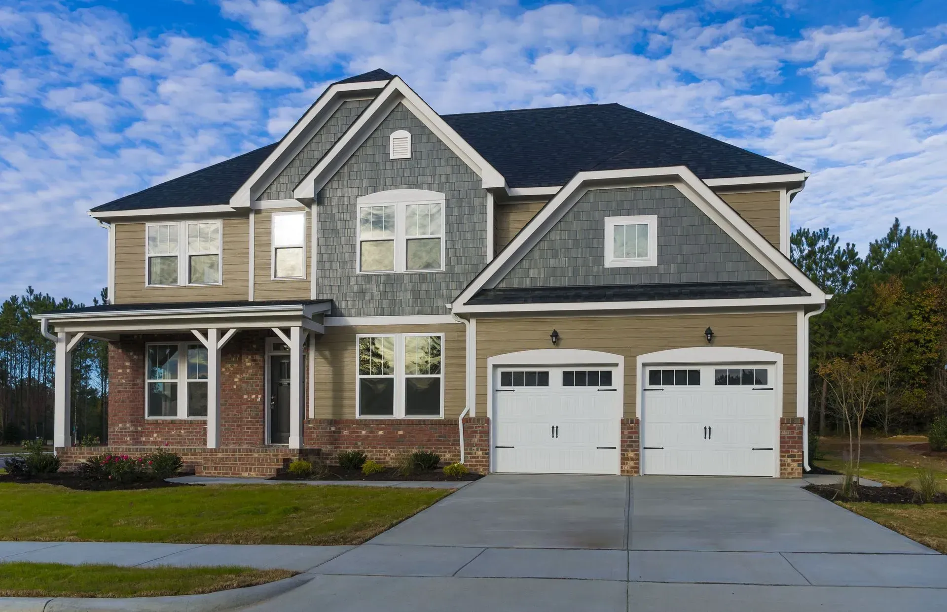 Two-story house with tan siding, brick accents, grey shingles, and a two-car garage under a blue sky with white clouds.
