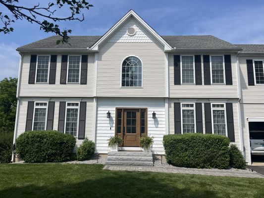 Two-story house with tan siding, black shutters, and brown front door; green bushes in front.
