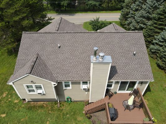 Aerial view of a house with a gray roof, chimney, deck, and surrounding greenery.
