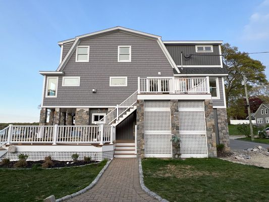 Gray house with deck, stairs, and stone supports, set against a blue sky.