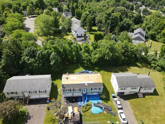 Aerial view of houses, one with roof repair in progress, surrounded by trees and green lawns.