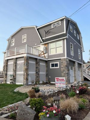 Gray and stone-faced house with porch overlooking a garden and lawn. Sign out front.
