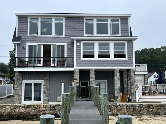 Two-story house with gray shingles, stone pillars, and a wooden dock on the water.