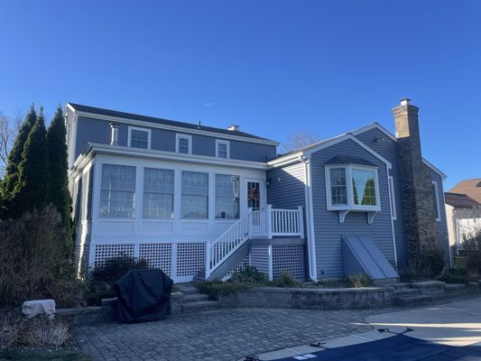 A light blue house with a stone chimney, sunroom, and white latticework on a clear day.