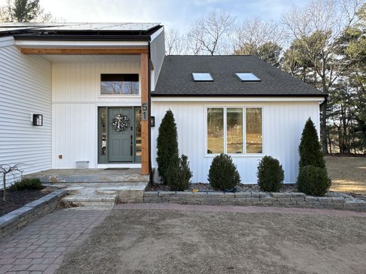 White house exterior with dark gray roof, green door, and small evergreen bushes in front.