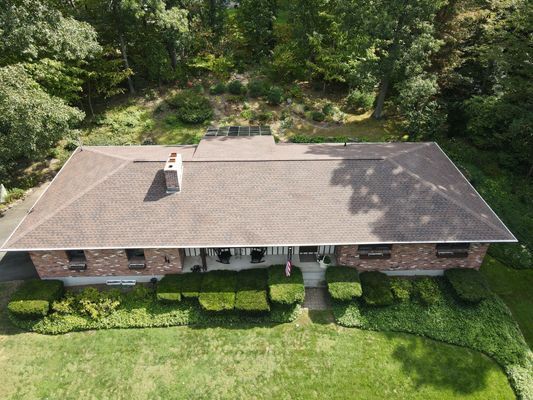 A brick house with a brown roof sits on a grassy hill surrounded by trees.