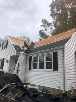 A house being re-roofed; a worker on the roof is installing shingles, surrounded by new wood and tarp.