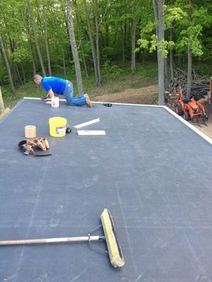 Person on hands and knees working on a dark gray flat roof with tools and a bucket. Trees and a tractor are in the background.