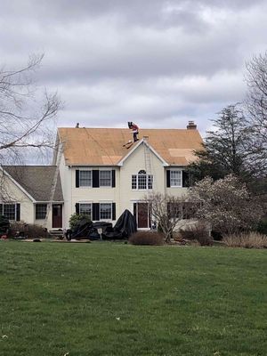 House with roofing being replaced; two workers on roof, brown shingles visible, black shutters, green lawn.