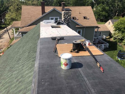 Rooftop with roofing tools, flat black surface, and green shingled side. Residential setting with houses in background.