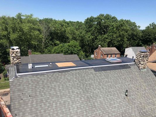 Overhead view of rooftops. Gray shingle roof, flat black roof with skylight, stone chimneys, trees in the background.