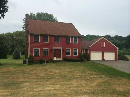Two-story red house with a brown roof and a two-car garage on a grassy lawn. Overcast sky.