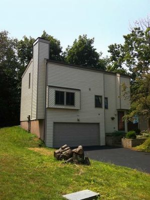 Beige two-story house with a garage and black driveway on a grassy hill, trees in the background.