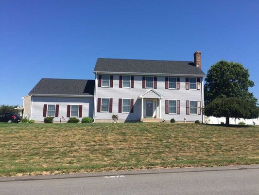 Two-story light gray house with dark shutters, a brick chimney, and a tree on a sunny day.