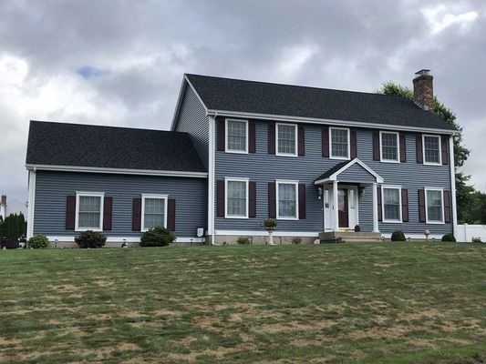 Two-story blue house with dark shutters and roof on a grassy hill under a cloudy sky.