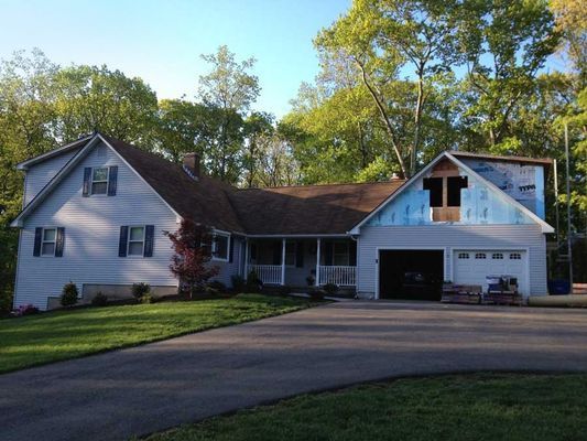 A light blue house with a dark driveway and a garage under construction. Trees in the background.