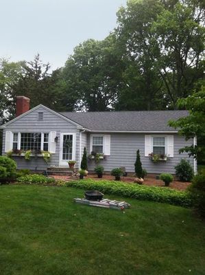 Gray-sided ranch house with white shutters, window boxes, brick path, and green lawn.
