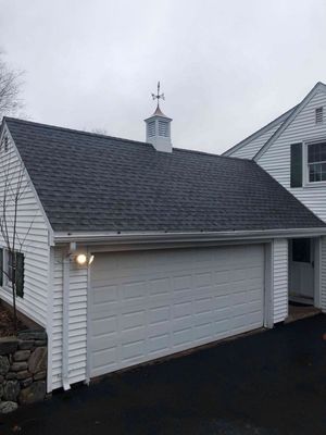 White garage with dark gray shingle roof, weather vane, and closed garage door, on an overcast day.