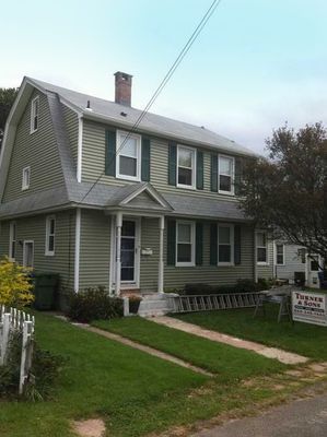 Green house with green shutters, a chimney, and a small porch on a sunny day.