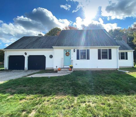 White house with dark roof and two-car garage. Blue door, wreath, and pumpkins on lawn. Sunny day.