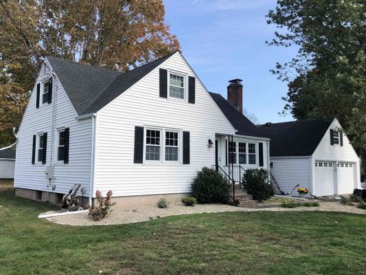White house with black shutters, dark roof, and attached garage on a grassy lot under a blue sky.