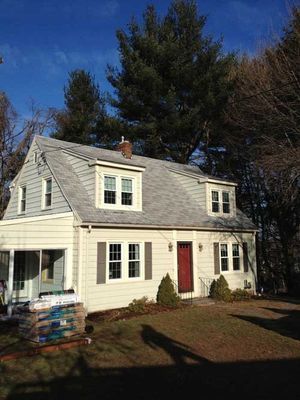 Tan house with grey roof, red door, and multiple windows; tall trees in the background under a blue sky.